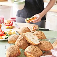 Ein aufgeschnittenes Weizenbrötchen mit heller, lockerer Krume und sieben goldgelb ausgebackene, mit Sesam bestreute Yoga Buns liegen auf einem Abkühlgitter.