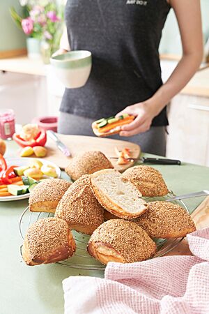 Ein aufgeschnittenes Weizenbrötchen mit heller, lockerer Krume und sieben goldgelb ausgebackene, mit Sesam bestreute Yoga Buns liegen auf einem Abkühlgitter.